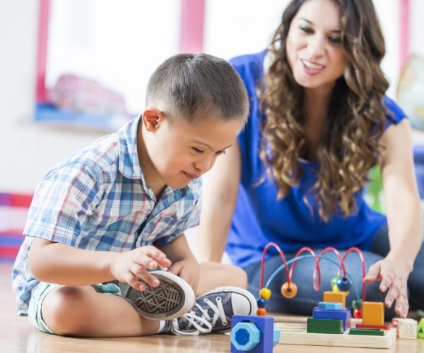 Sweet little Hispanic boy with Down Syndrome sits cross legged on the floor of a daycare center and reaches for toys. His female Hispanic young adult teacher sits on the floor with him and reaches for a different toy.